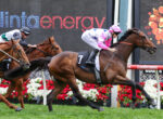 Two jockeys race brown horses toward the finish, pink-and-white-silk rider ahead as flowers line the track and a digital display looms in the background.