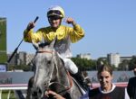 Jockey in yellow silks on a gray horse celebrates victory, raising a fist as a handler holds the reins nearby at a sunny racetrack.