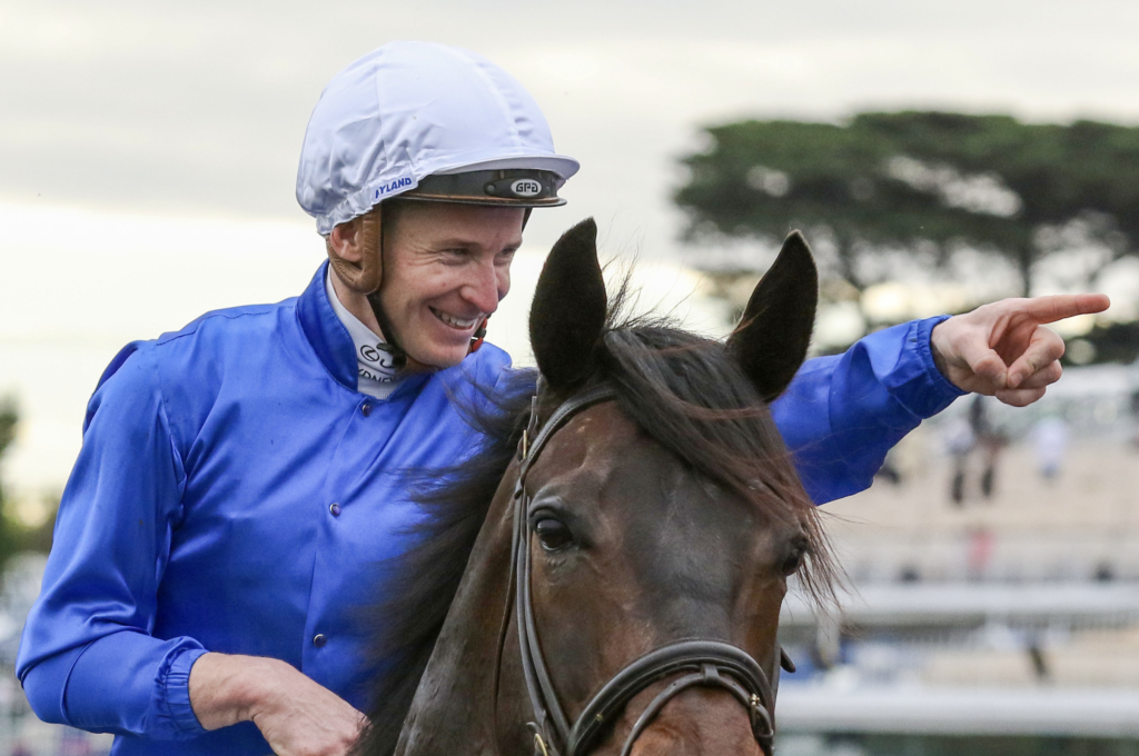 Jockey wearing a blue jacket and white helmet rides a brown horse, smiling and pointing to the right as they move past.