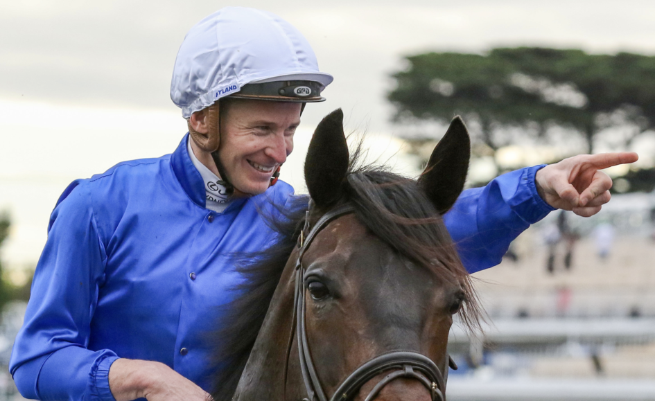 Jockey wearing a blue jacket and white helmet rides a brown horse, smiling and pointing to the right as they move past.