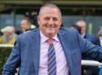Smiling man in a blue plaid suit and pink shirt at an outdoor event, wearing a colorful striped tie.