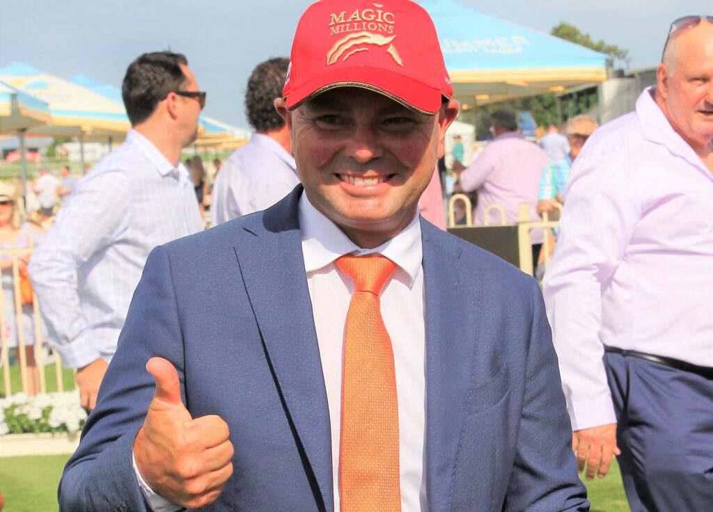 Smiling man in a navy suit and orange tie giving a thumbs-up at an outdoor event, wearing a red cap that says 'Magic Millions'