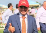 Smiling man in a navy suit and orange tie giving a thumbs-up at an outdoor event, wearing a red cap that says 'Magic Millions'