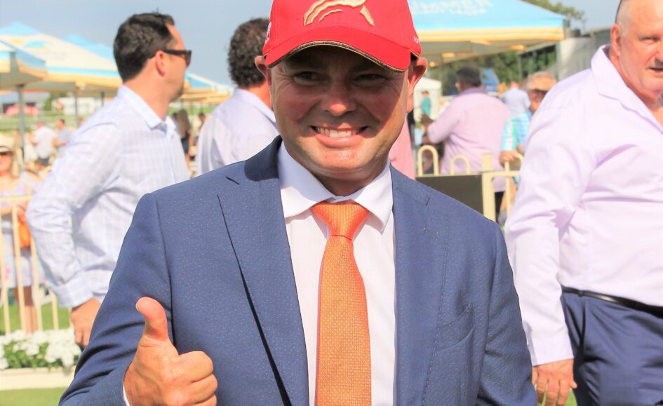 Smiling man in a navy suit and orange tie giving a thumbs-up at an outdoor event, wearing a red cap that says 'Magic Millions'