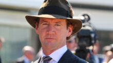 Man in a brown fedora, pinstripe suit, and patterned tie at a formal outdoor event, facing the camera.