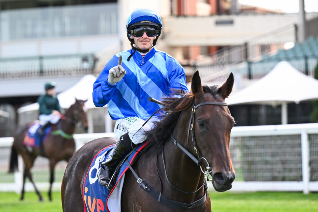 Jockey in blue and white silks on a brown horse, wearing helmet and goggles, raising one finger toward the camera during a race.