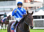 Jockey in blue and white silks on a brown horse, wearing helmet and goggles, raising one finger toward the camera during a race.