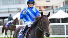 Jockey in blue and white silks on a brown horse, wearing helmet and goggles, raising one finger toward the camera during a race.