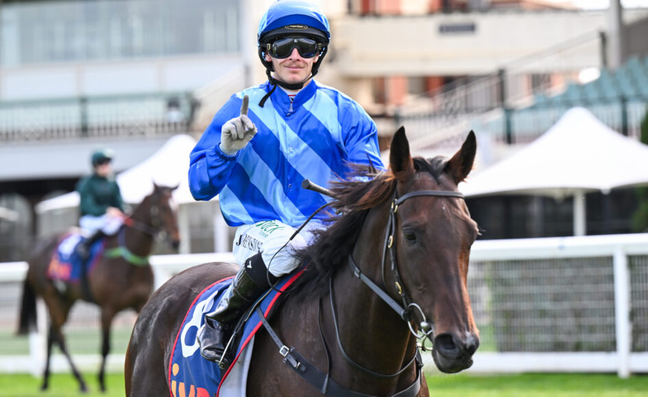 Jockey in blue and white silks on a brown horse, wearing helmet and goggles, raising one finger toward the camera during a race.