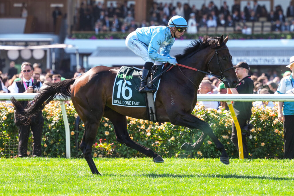 Jockey in light blue silks rides a dark horse with a green saddle cloth showing number 16 during a race, crowd in stands in the background.