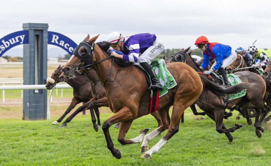 Jockey in purple and white leads a tight horse race on a green turf track with other riders close behind in colorful silks.