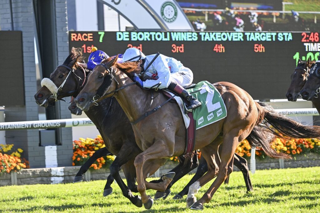 Three jockeys battle for the lead in a horse race, with a digital scoreboard behind them showing race details.