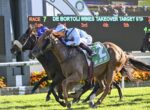 Three jockeys battle for the lead in a horse race, with a digital scoreboard behind them showing race details.