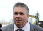 Close-up of a man in a gray suit and striped tie at an outdoor event with a blurred crowd in the background