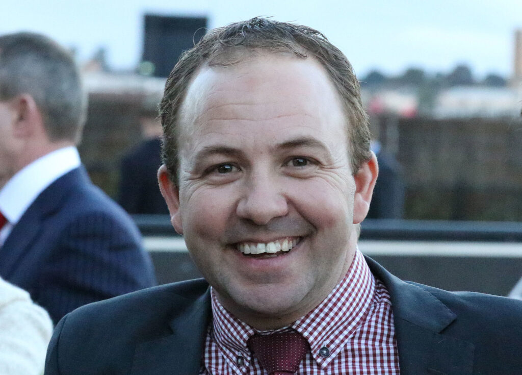 Close-up of smiling man in a navy suit and red checkered shirt at an outdoor event, blurred background.