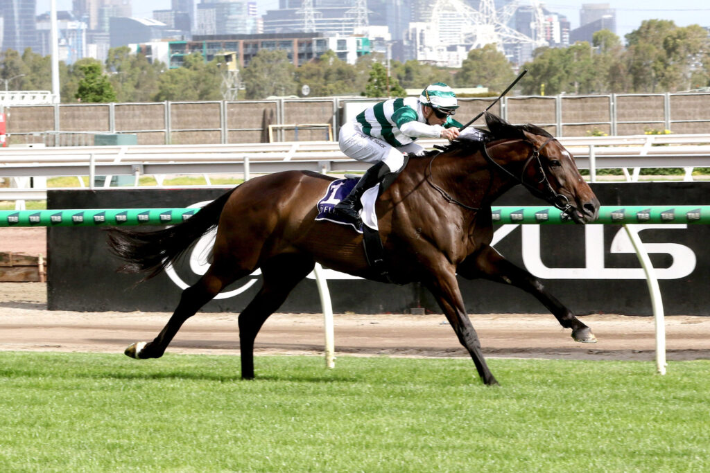 Horse and jockey in green-and-white silks race along a dirt track with a city skyline in the background.