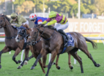 Three racehorses with jockeys sprint toward the finish on a sunny turf track, rails in the background.]?