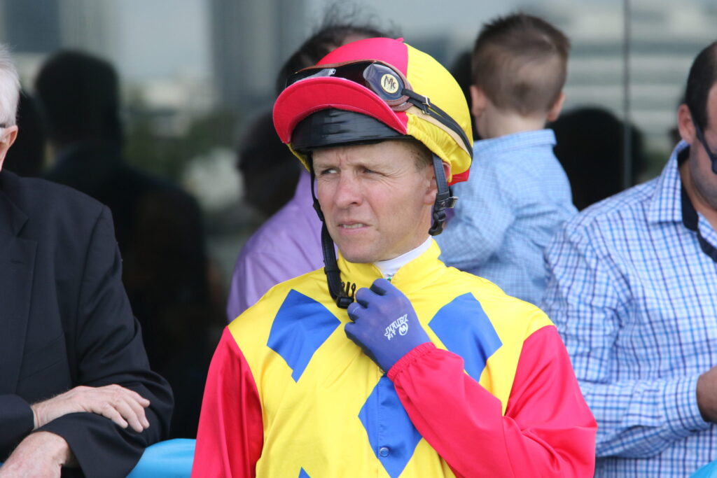 Man in a bright yellow, red, and blue racing suit adjusts a red helmet with goggles on top near others in background.