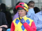 Man in a bright yellow, red, and blue racing suit adjusts a red helmet with goggles on top near others in background.