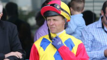 Man in a bright yellow, red, and blue racing suit adjusts a red helmet with goggles on top near others in background.