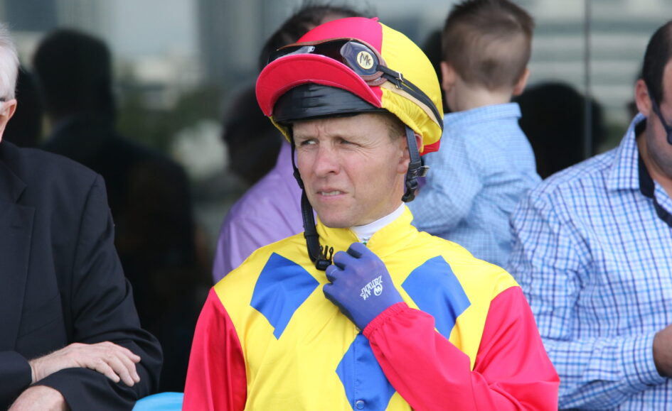 Man in a bright yellow, red, and blue racing suit adjusts a red helmet with goggles on top near others in background.