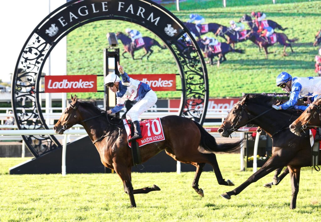 Two jockeys race on thoroughbreds near a decorative arch at Eagle Farm, with Ladbrokes banners in the background.