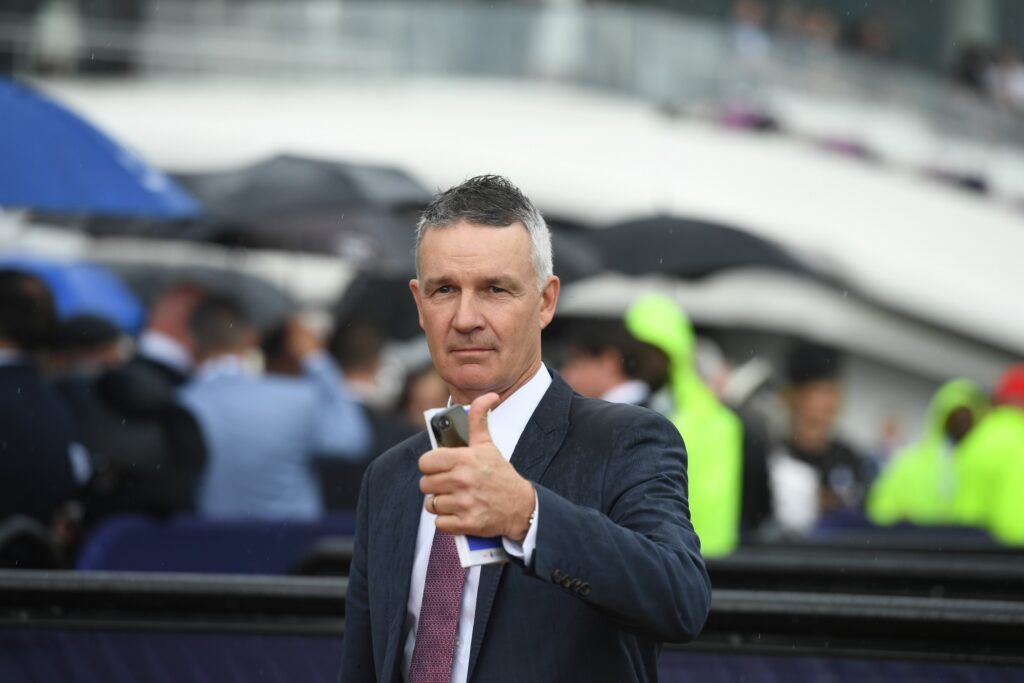 Man in a dark suit and red tie gives a thumbs-up at a crowded outdoor event in the rain, holding a smartphone and papers.