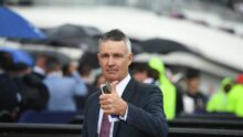 Man in a dark suit and red tie gives a thumbs-up at a crowded outdoor event in the rain, holding a smartphone and papers.