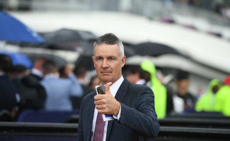 Man in a dark suit and red tie gives a thumbs-up at a crowded outdoor event in the rain, holding a smartphone and papers.