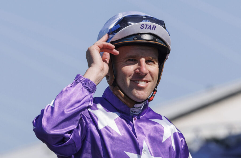 Jockey in purple star-patterned racing silks adjusting a helmet outdoors, smiling at the camera, blue sky behind.