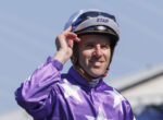 Jockey in purple star-patterned racing silks adjusting a helmet outdoors, smiling at the camera, blue sky behind.