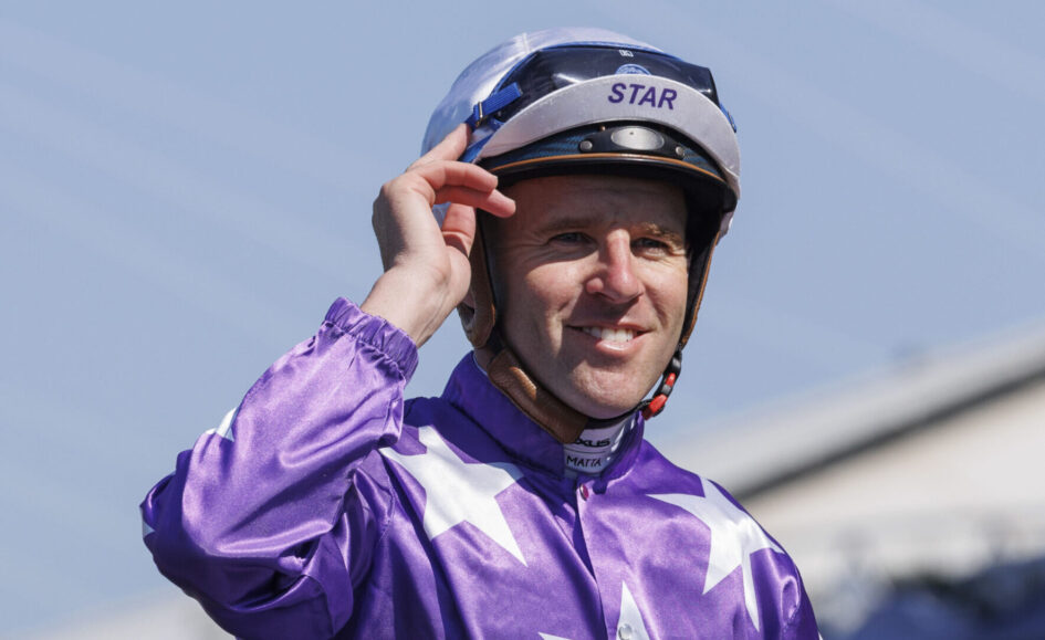 Jockey in purple star-patterned racing silks adjusting a helmet outdoors, smiling at the camera, blue sky behind.