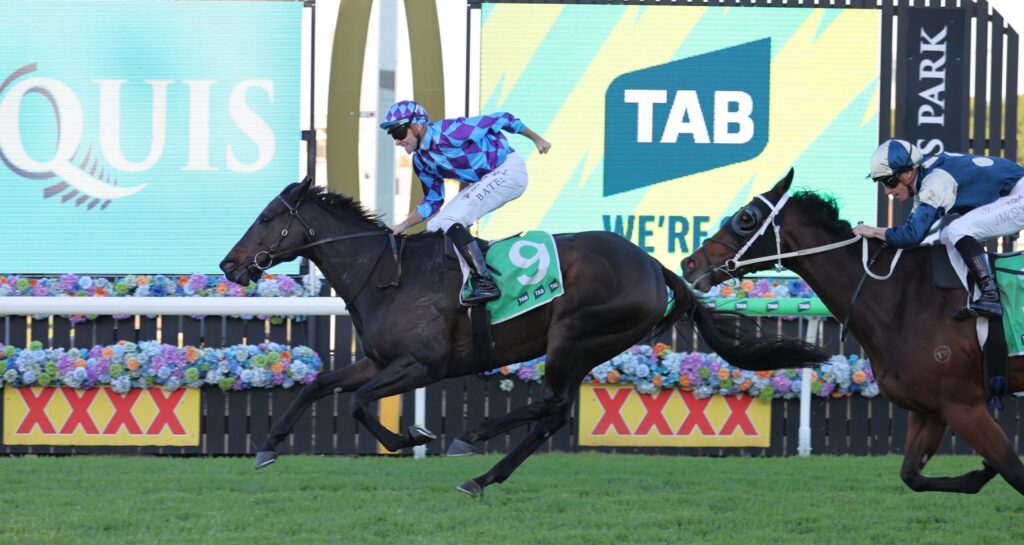 Two jockeys sprint toward the finish on galloping horses, the leading horse wearing a green saddlecloth with number 9; bright screens and flower barriers rise in the background.