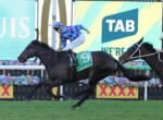 Two jockeys sprint toward the finish on galloping horses, the leading horse wearing a green saddlecloth with number 9; bright screens and flower barriers rise in the background.