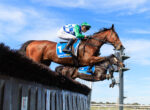 Jockey in green and blue silks rides a brown horse mid-jump over a track barrier against a blue sky.