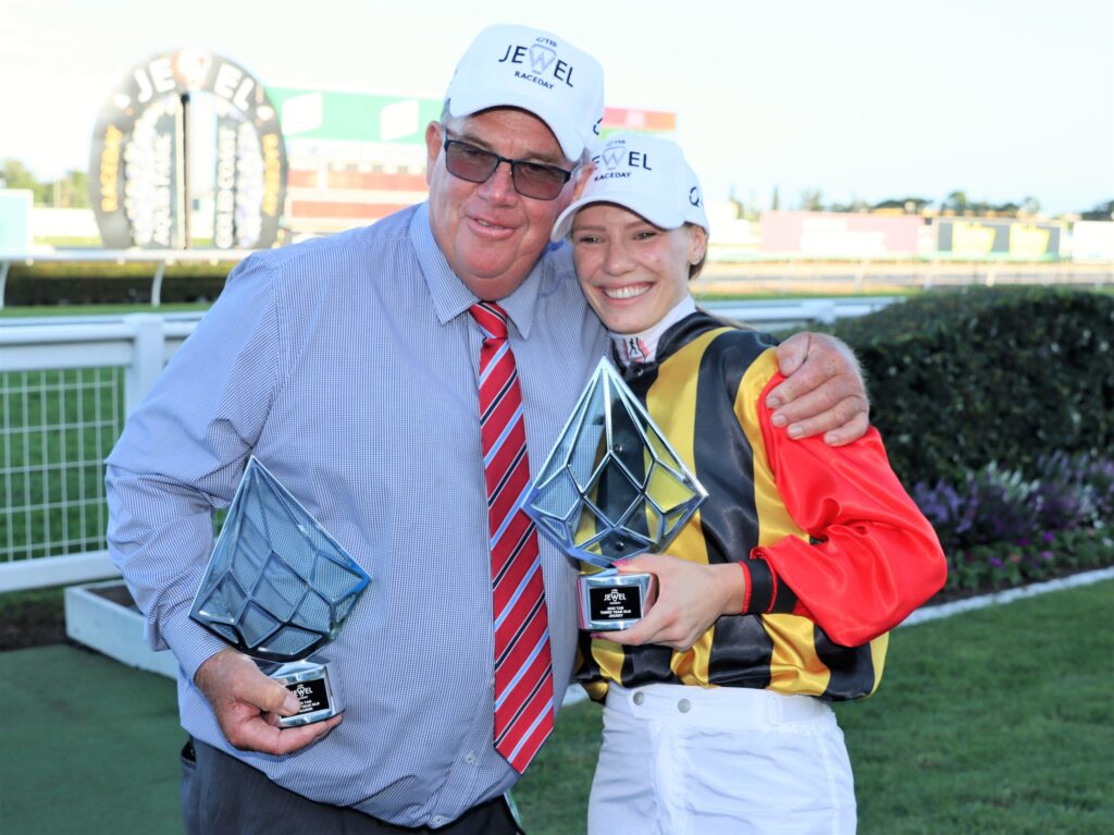 Two people pose with tall geometric trophies after a race event, smiling at the camera outdoors with a fence in the background.