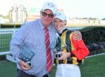 Two people pose with tall geometric trophies after a race event, smiling at the camera outdoors with a fence in the background.