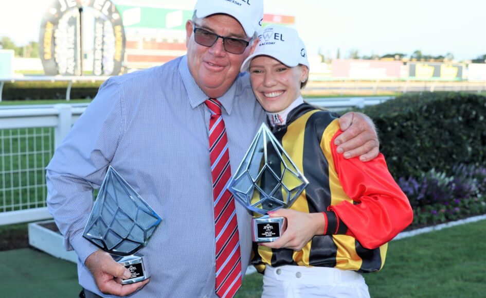 Two people pose with tall geometric trophies after a race event, smiling at the camera outdoors with a fence in the background.