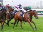 Jockeys in colorful silks race at full speed on a wet, grassy track with splashing water droplets.