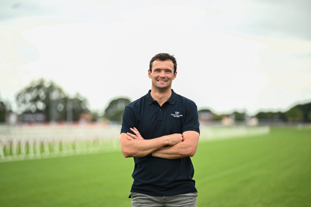 Man standing on a grassy sports field with arms crossed, wearing a dark polo shirt with a small chest logo.