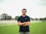 Man standing on a grassy sports field with arms crossed, wearing a dark polo shirt with a small chest logo.