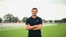 Man standing on a grassy sports field with arms crossed, wearing a dark polo shirt with a small chest logo.