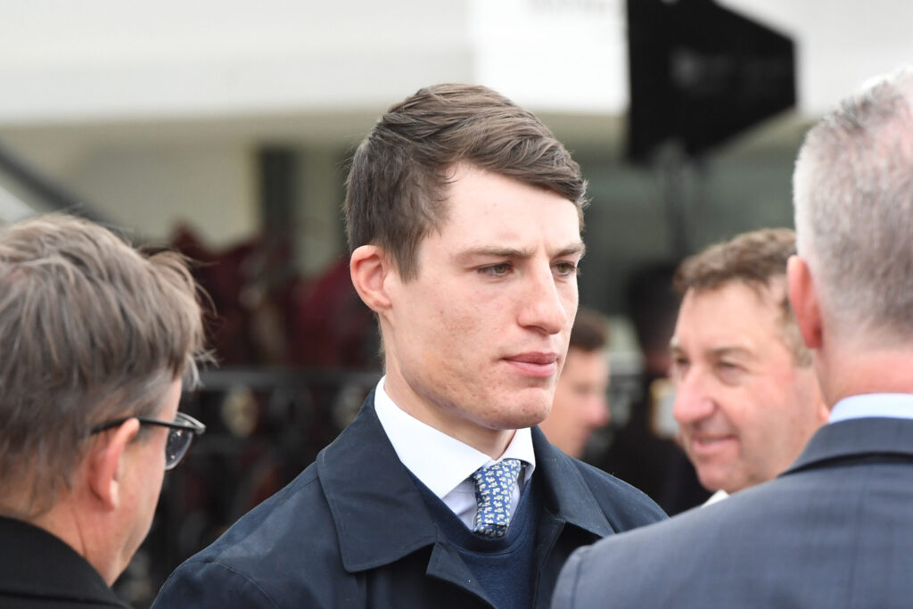 Young man in a navy suit and patterned tie talking at a formal event, surrounded by people in the background.