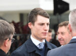 Young man in a navy suit and patterned tie talking at a formal event, surrounded by people in the background.