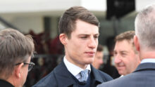 Young man in a navy suit and patterned tie talking at a formal event, surrounded by people in the background.