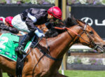 Jockey in red helmet leaning forward on a brown horse during a race, green saddlecloth with TAB logos visible and a second horse nearby.