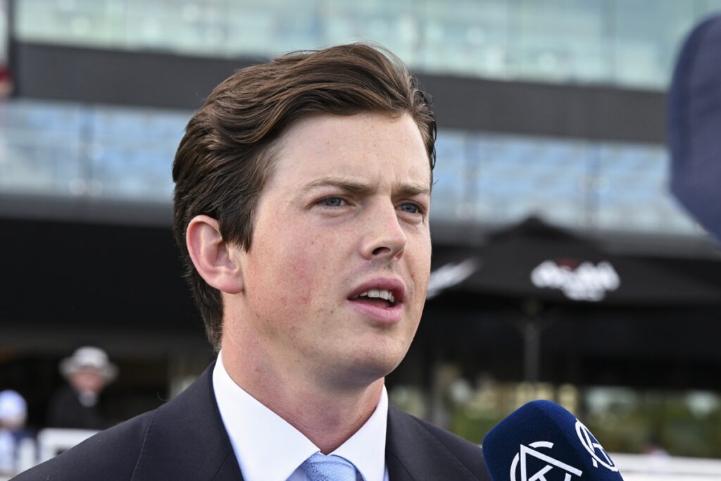Man in a dark suit and light blue tie speaks into a blue microphone during an outdoor interview