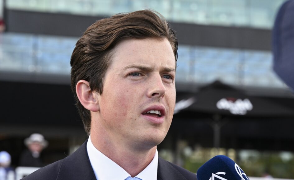 Man in a dark suit and light blue tie speaks into a blue microphone during an outdoor interview