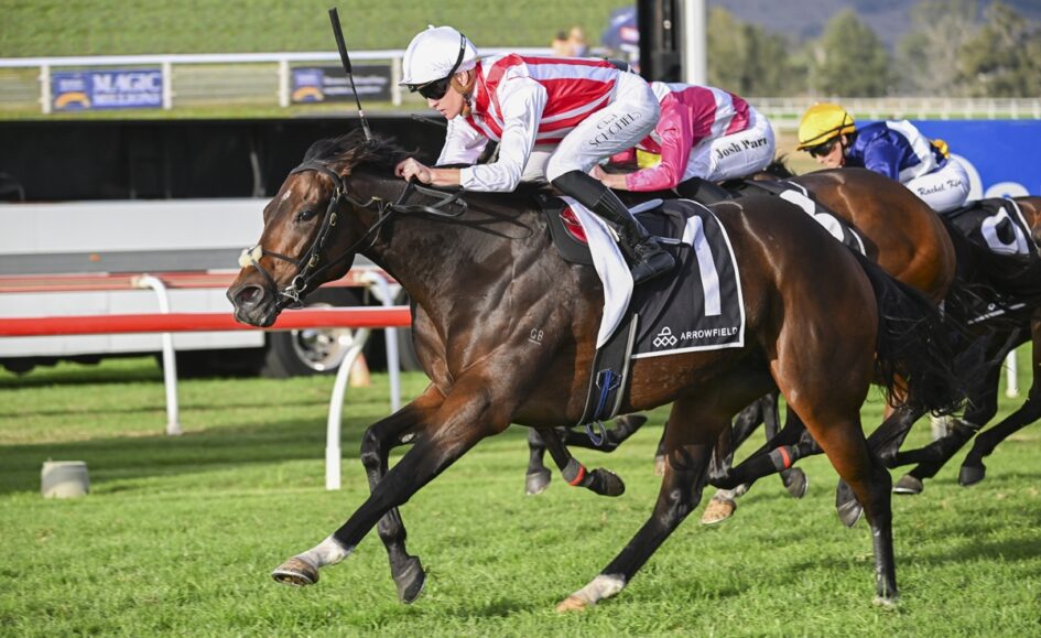 Dark brown racehorse and rider in red‑and‑white silks sprint on a turf track, first past the rails in a crowded race.