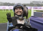Jockey in muddy blue riding gear and helmet smiling at the camera, holding up three fingers at a racecourse with a horse in the background.